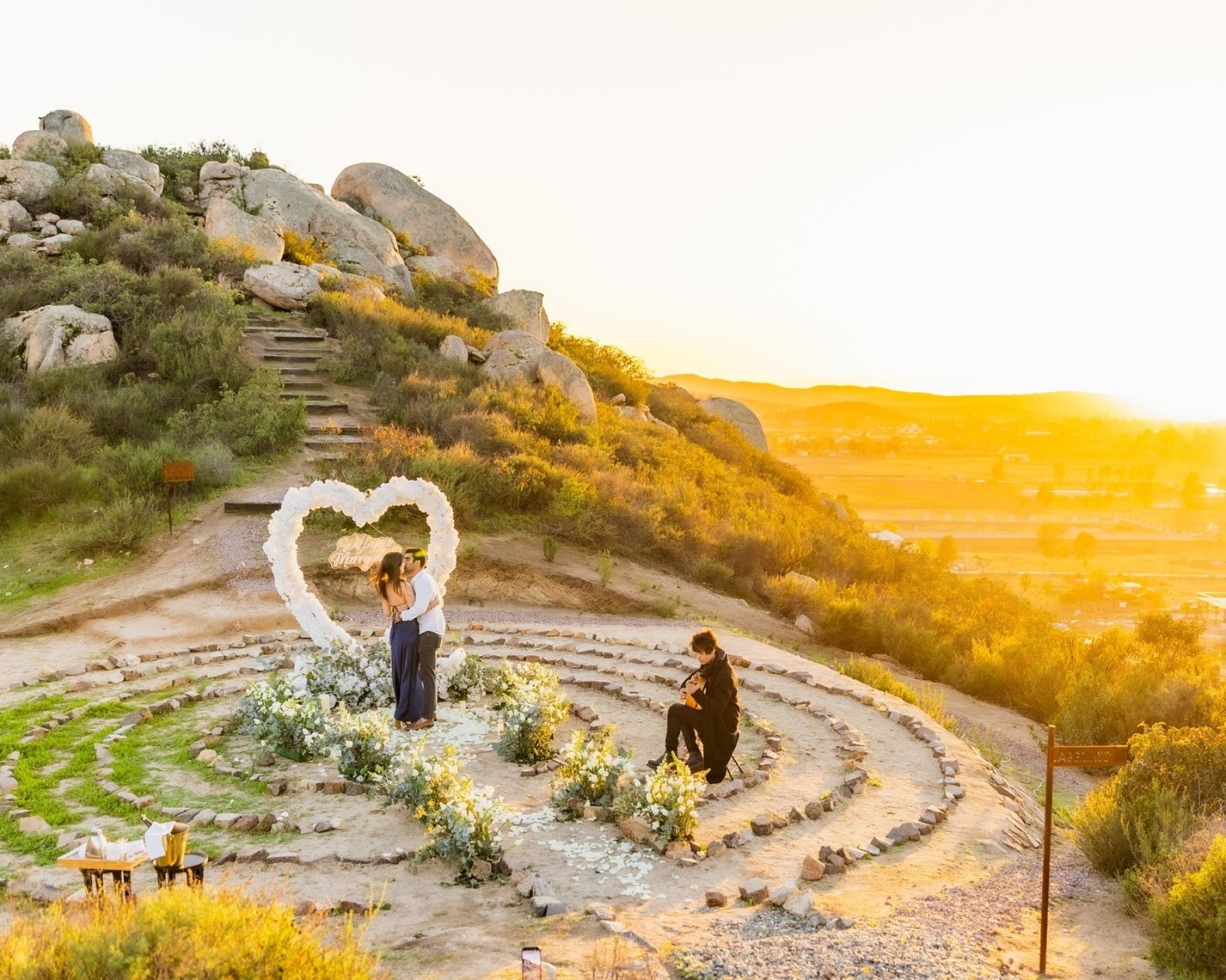 The main proposal photo: Brij proposing to Cate among concentric stone steps, flowers, and a heart-shaped arch at sunset